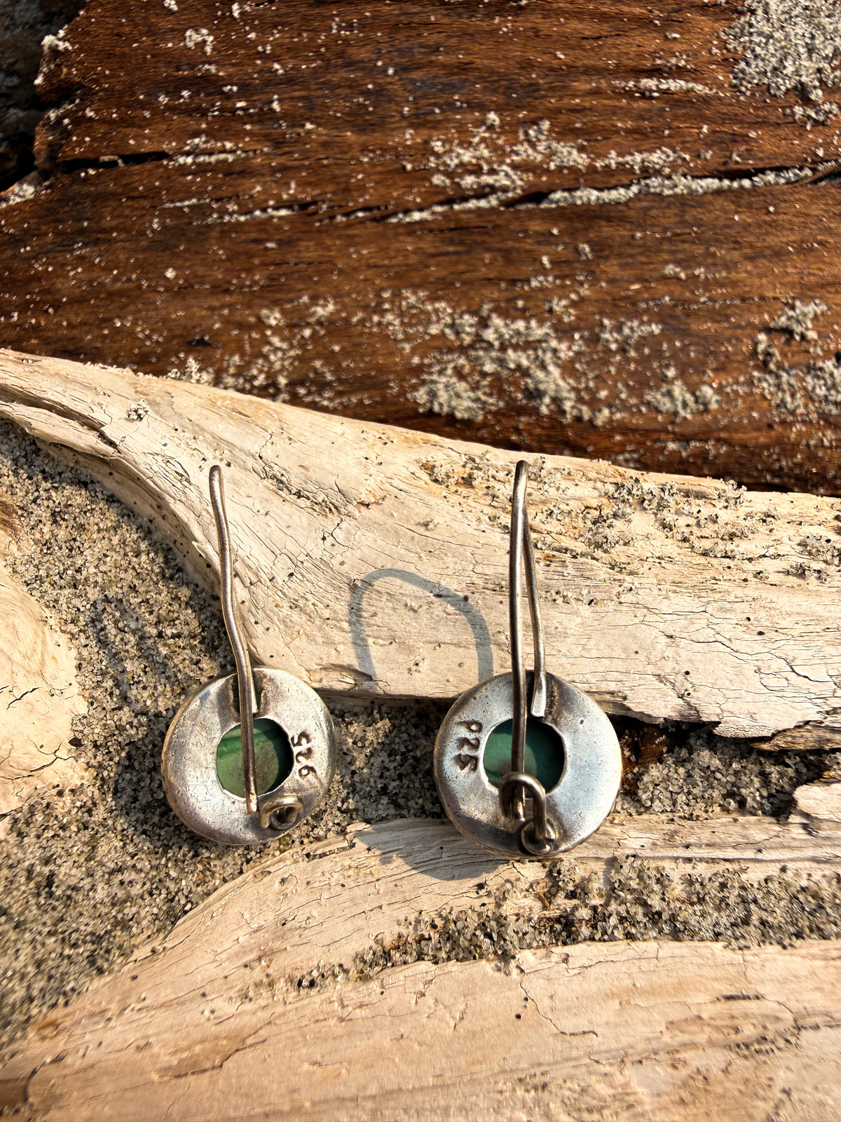 A pair of handmade sterling silver earrings with circular disc pendants featuring a green stone inlay, displayed on a piece of driftwood over sand.