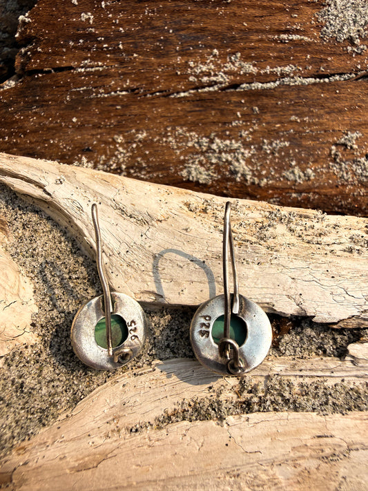 A pair of handmade sterling silver earrings with circular disc pendants featuring a green stone inlay, displayed on a piece of driftwood over sand.